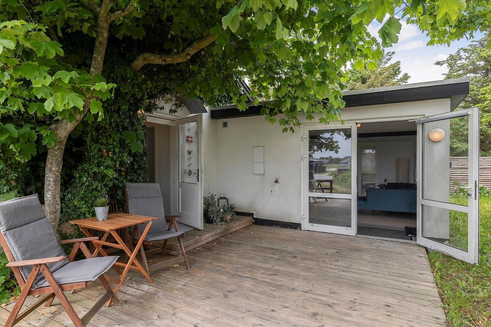 Terrasse mit Tisch und Stühlen unter einem großen Baum. Eingang und Schiebetür führen ins Haus.