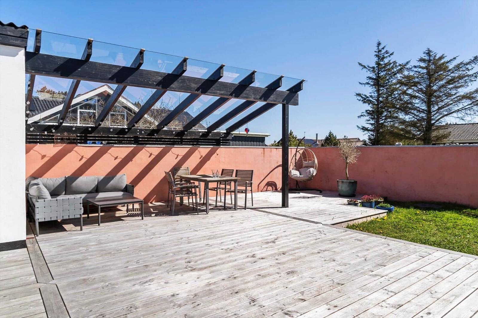 Terrasse mit Sitzgruppe, Tisch, Hängesessel und Glasdach. Hinterhof mit rosa Wand und Grünfläche.