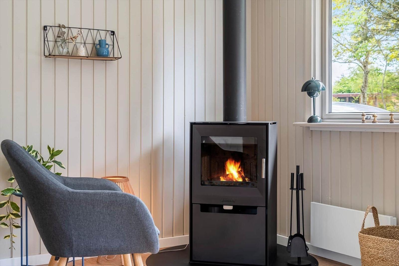 Kaminofen mit brennendem Feuer, graue Sitzgelegenheit, Fenster mit Blick auf Grün