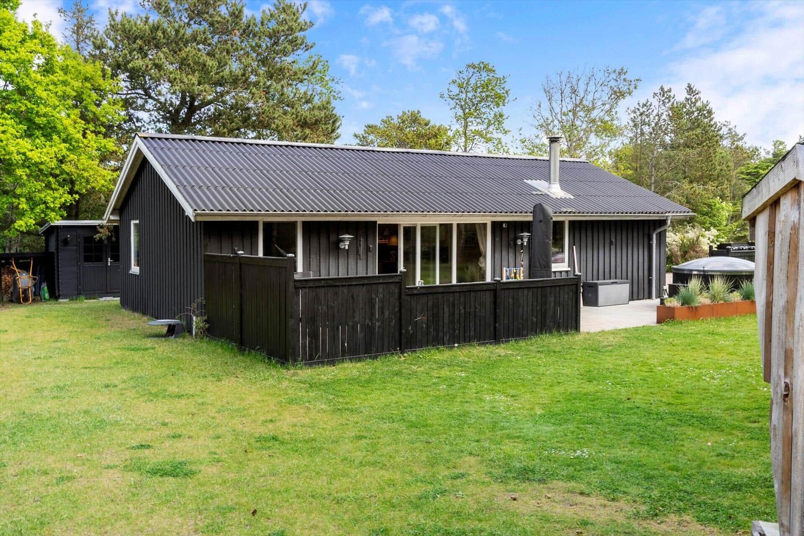 Ein dunkelgefärbtes Holzhaus mit Balkon und Garten. Im Hintergrund sind Bäume sichtbar.