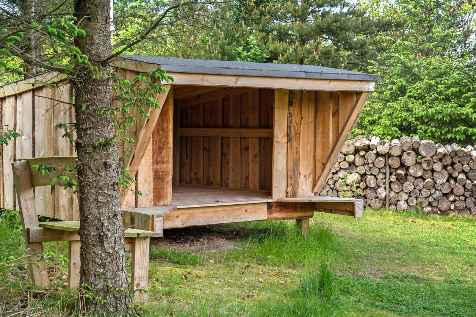 Holzkonstruktion mit Dach, umgeben von Bäumen und einem Holzstapel.
