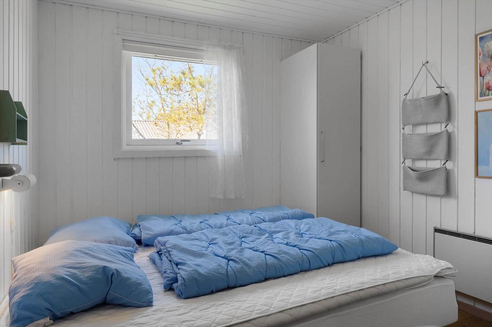 Schlafzimmer mit Bett, Fenster und Kleiderschrank an weißen Wänden.