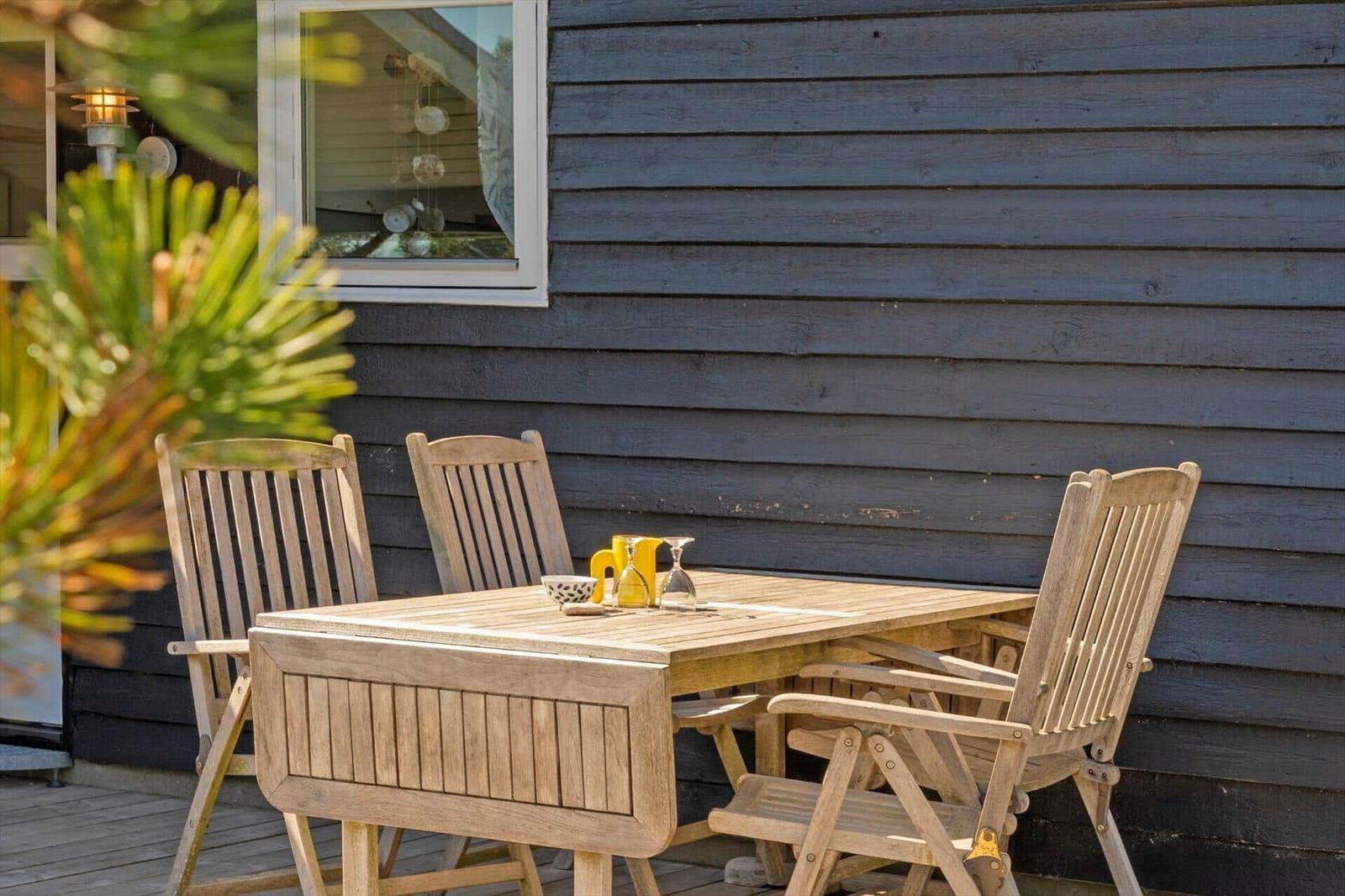 Holztisch mit Stühlen auf Terrasse vor grauem Holzhaus mit Fenster.