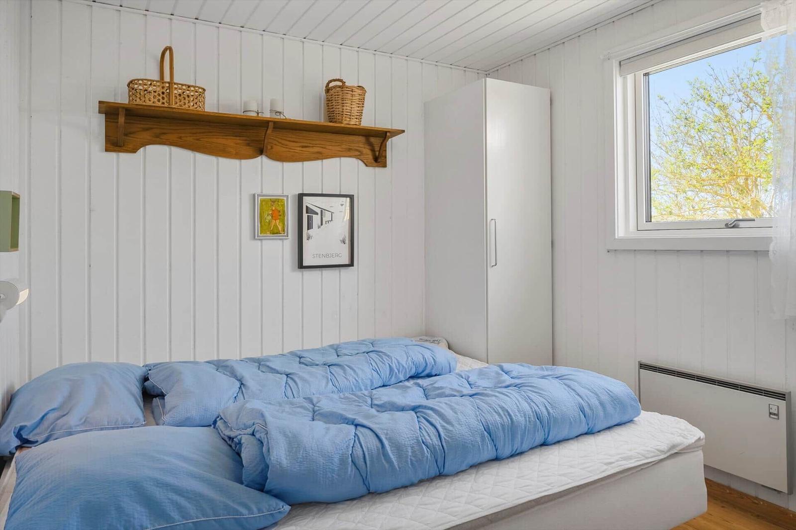 Schlafzimmer mit blauer Bettwäsche, weißer Wandverkleidung und Fenster mit Blick auf Bäume.