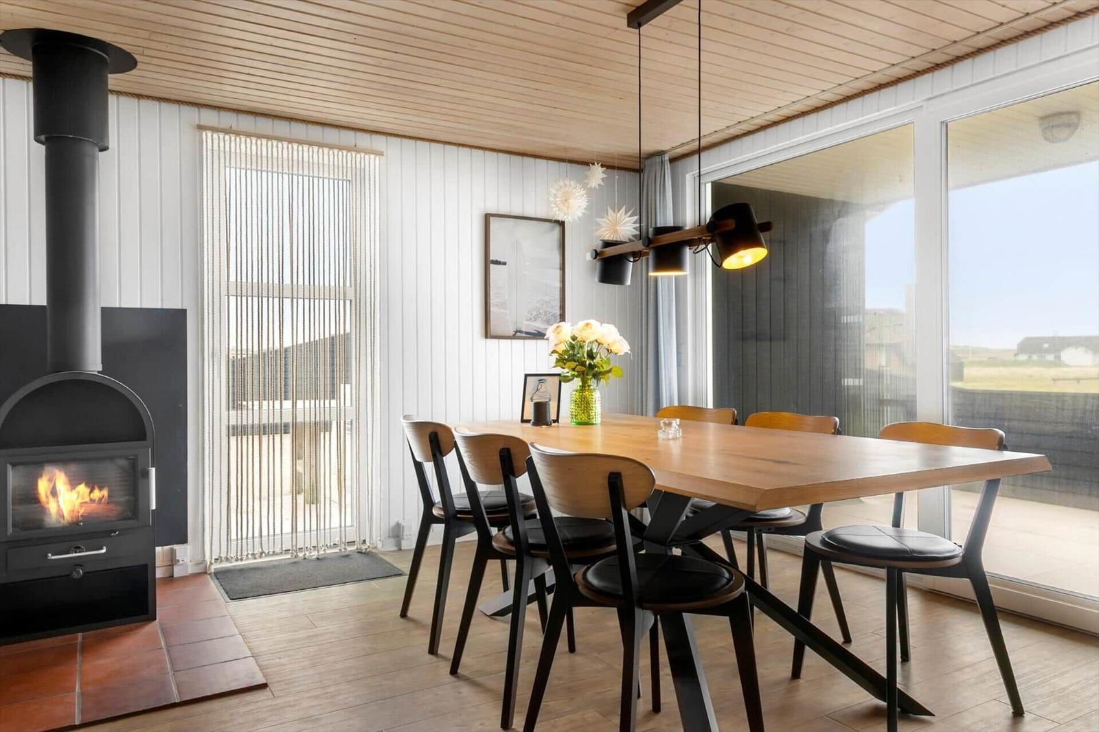 Dining room with wooden table, chairs, and wood stove. Large windows and ceiling paneling.