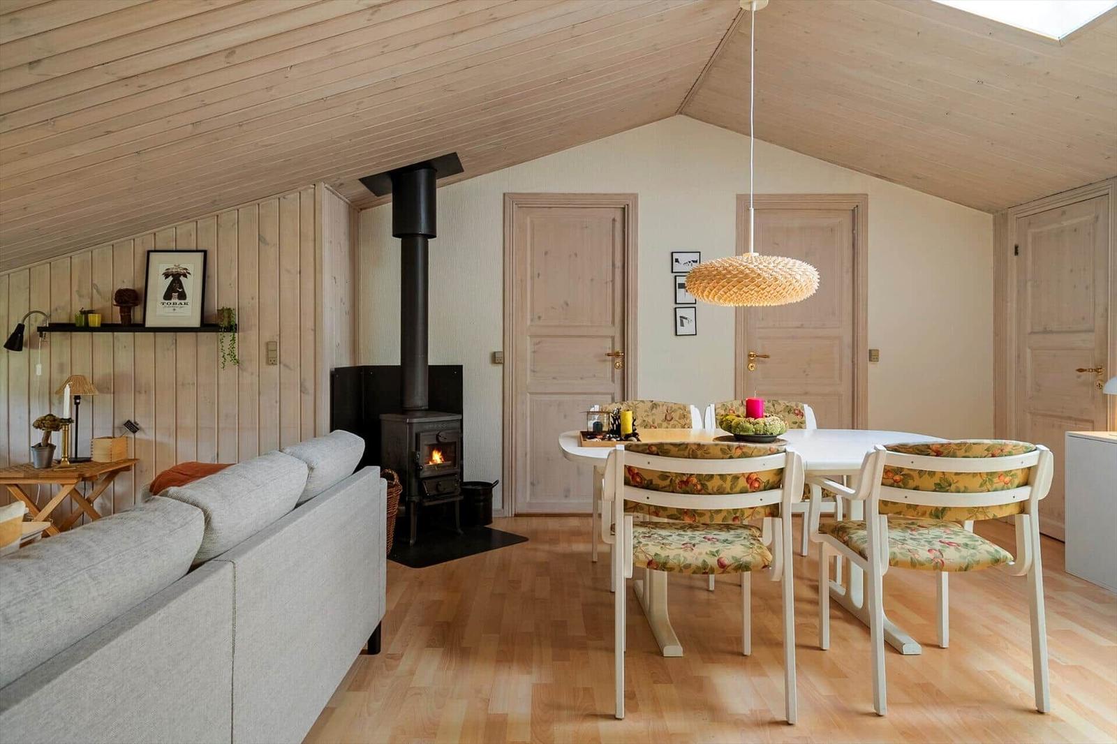 Wohn- und Essbereich mit Holzdecke, Kamin und Stuhlgruppe mit floraler Polsterung.