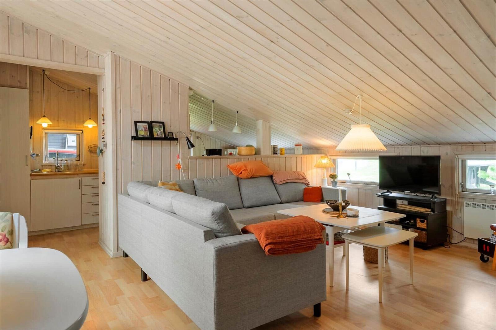 Wohnzimmer mit Sofa, Tisch und TV. Küche im Hintergrund. Holzdecke und -wände.