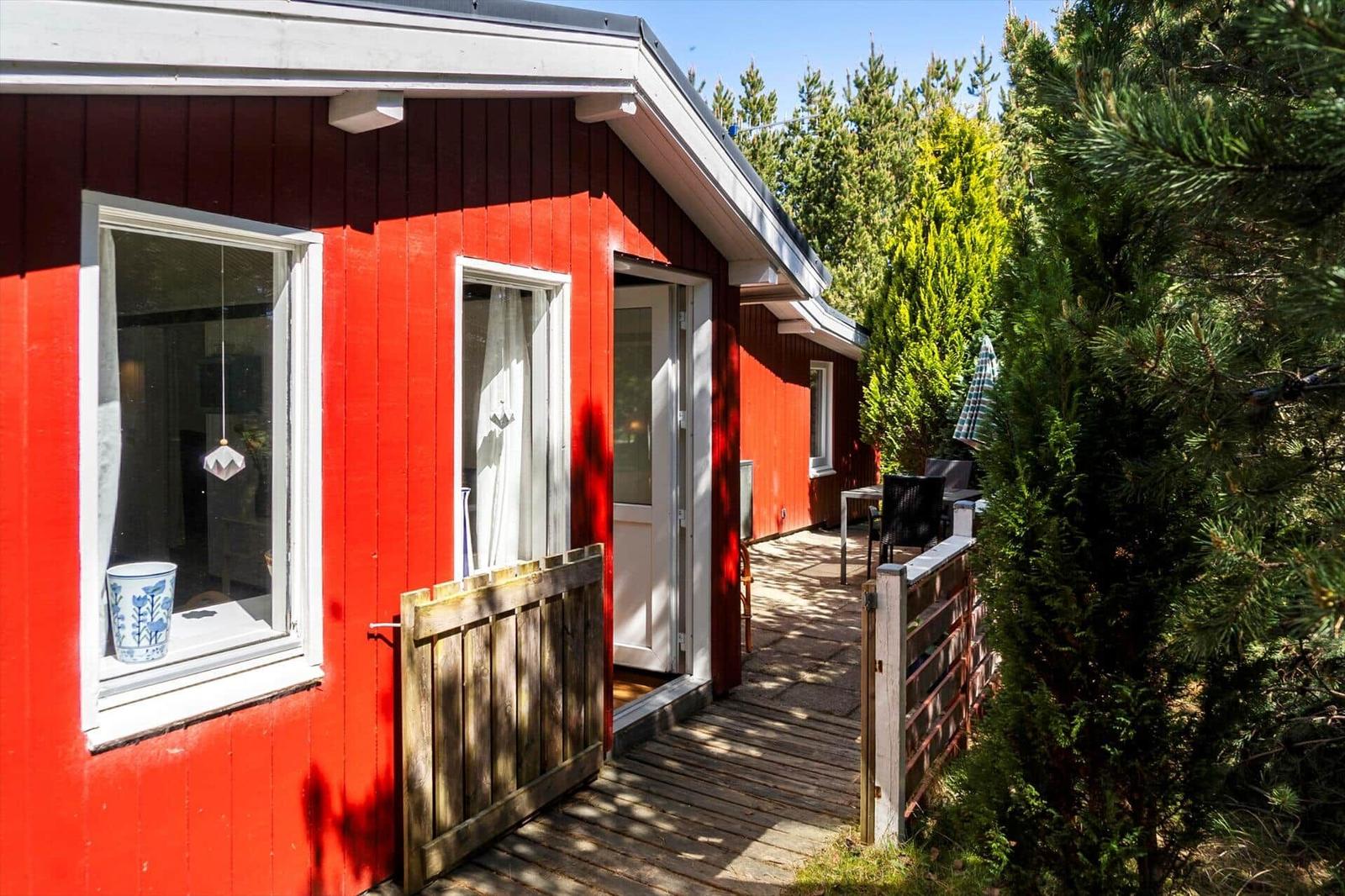 Rotes Holzhaus mit Terrasse und Fenstern. Holztor und Bäume sind sichtbar.