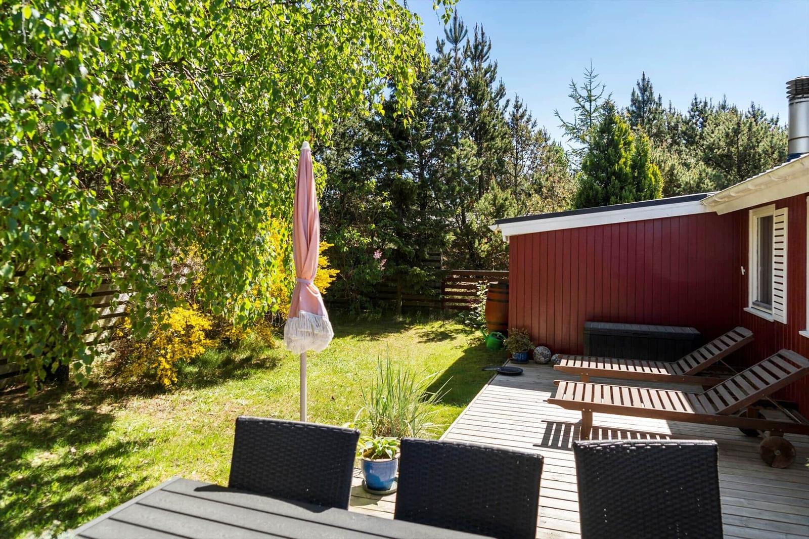 Terrasse mit Tisch, Stühlen und Sonnenliegen neben rotem Haus. Grünfläche und Bäume im Hintergrund.