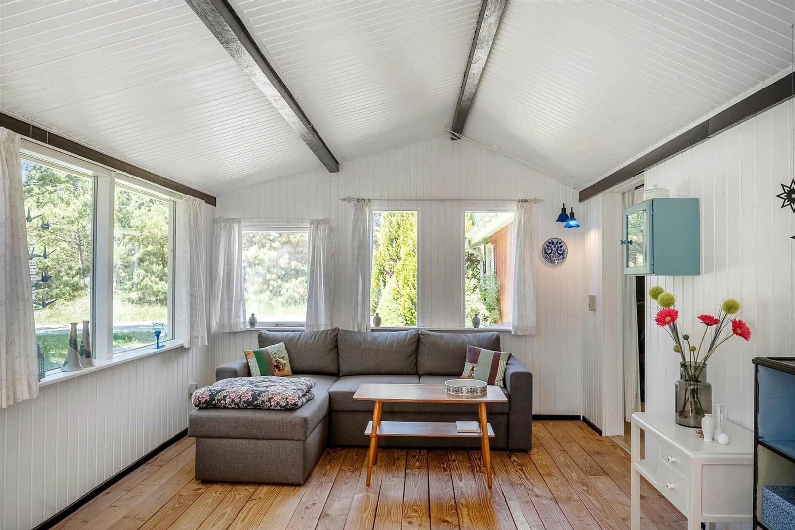Ein Wohnzimmer mit grauem Sofa, Holzboden und weißen Wänden. Große Fenster mit Blick auf Grün.