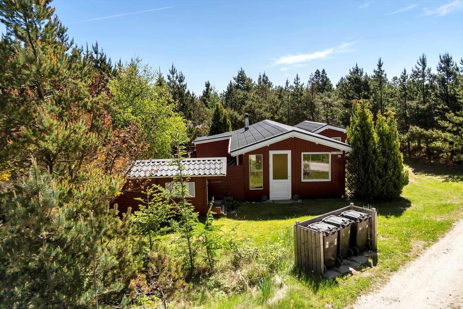 Rotes Holzhaus mit weißen Fensterrahmen und Dach im Wald. Grasfläche und Weg führen zum Eingang.