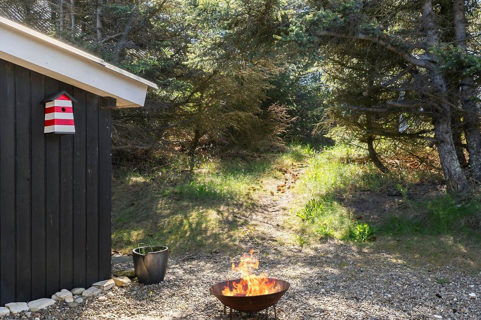Außenbereich mit Feuerkorb, dunklem Holzhaus und rotem Streifen-Vogelhaus.