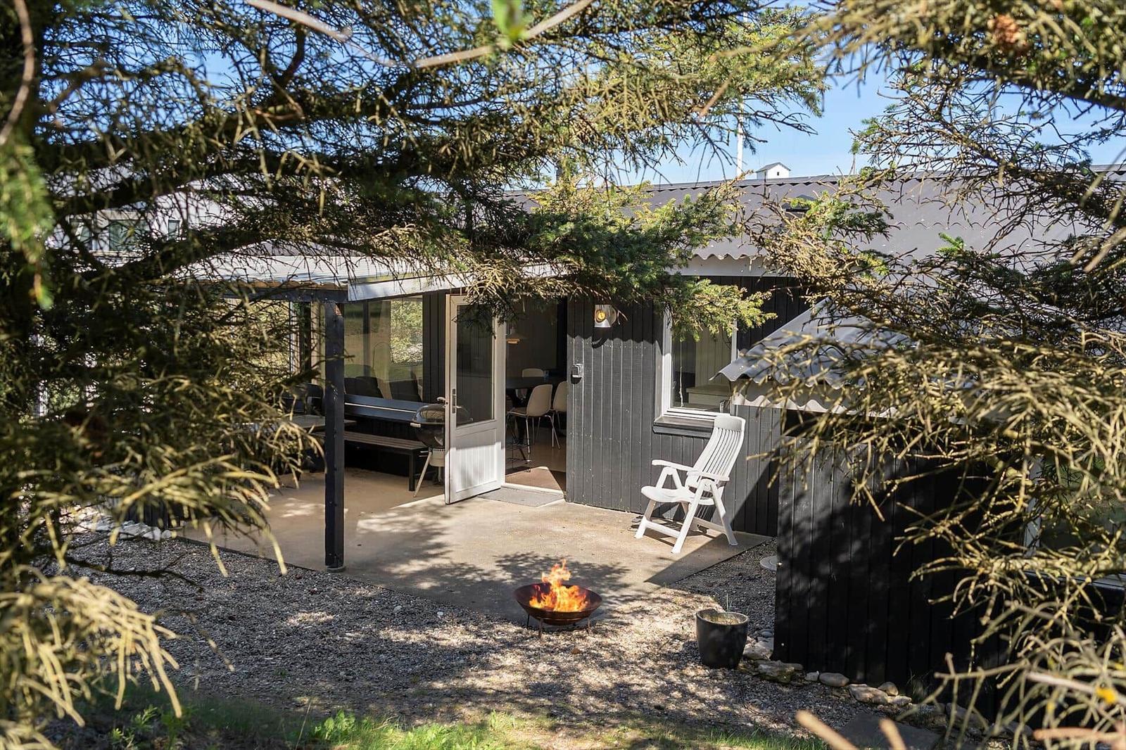 Hütte mit Terrasse, Sitzgelegenheit und brennendem Feuerkorb unter Bäumen.