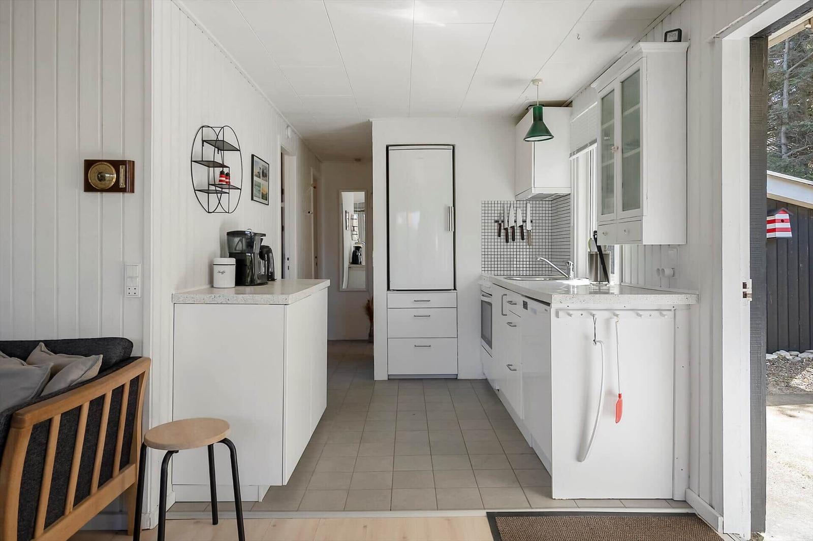 Küche mit weißen Schränken, Kühlschrank und Arbeitsplatte. Links Wohnbereich mit Sofa und Stuhl.