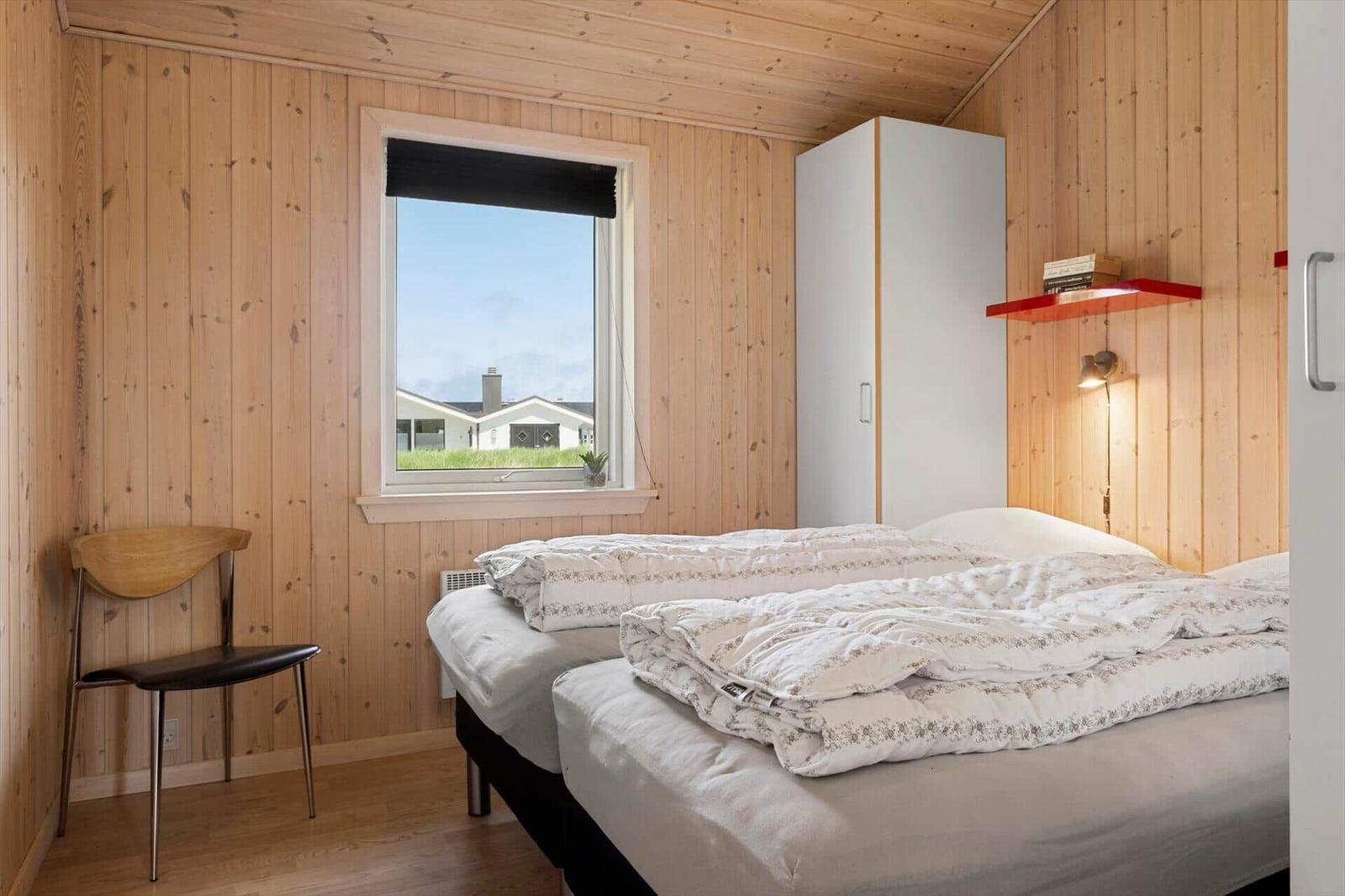 Schlafzimmer mit Holzwand, Bett, Stuhl und Fensterblick.