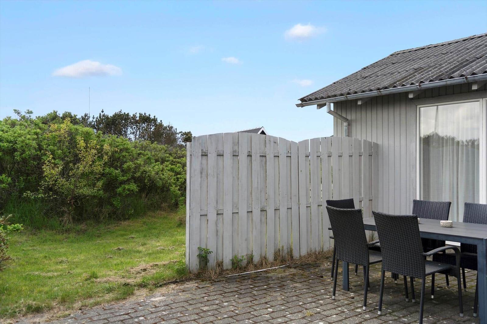 Terrace with table and chairs next to a house with white fence and green area.
