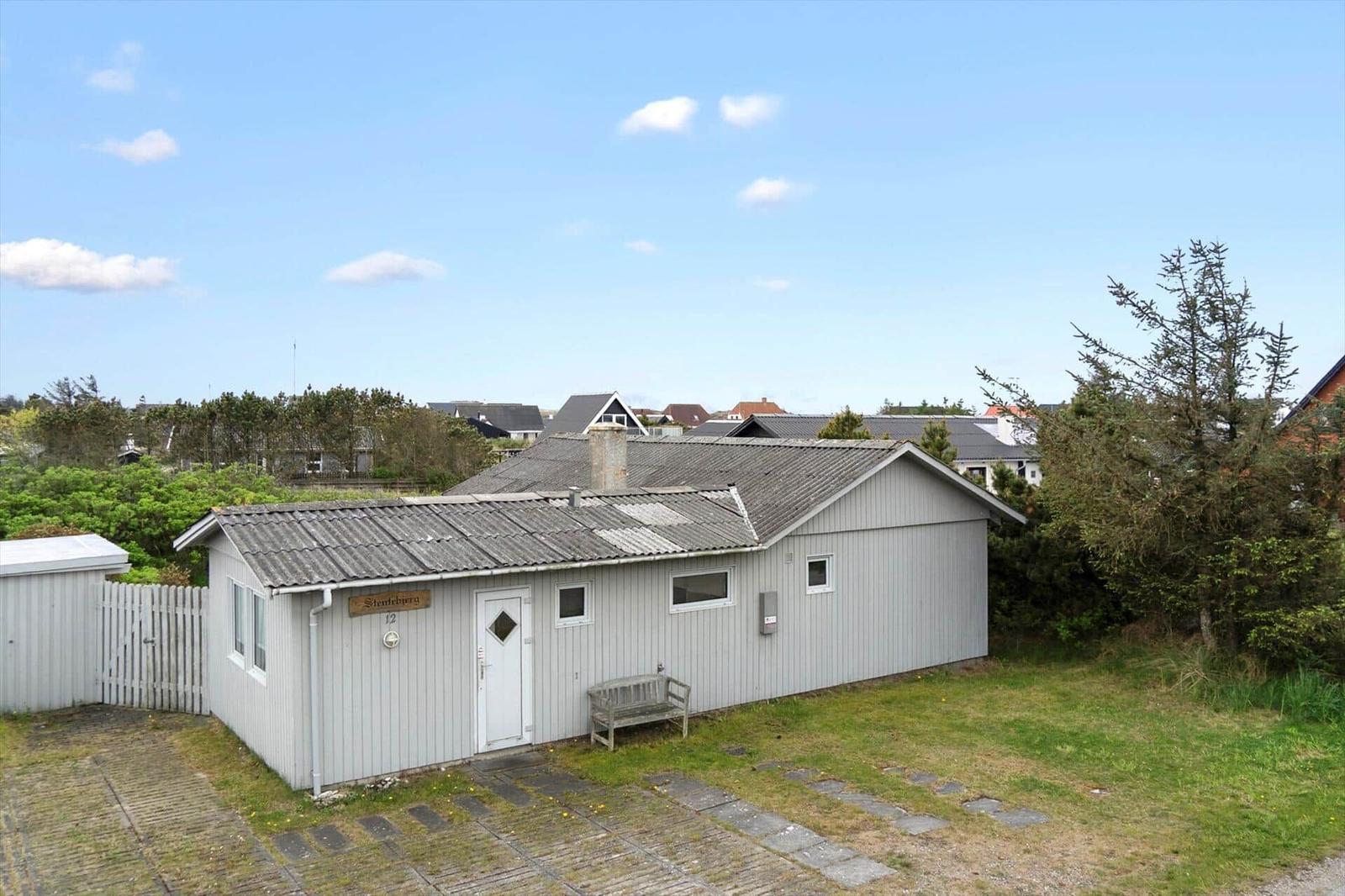 A small house with gray siding and a front yard. A white door and a wooden bench are visible.