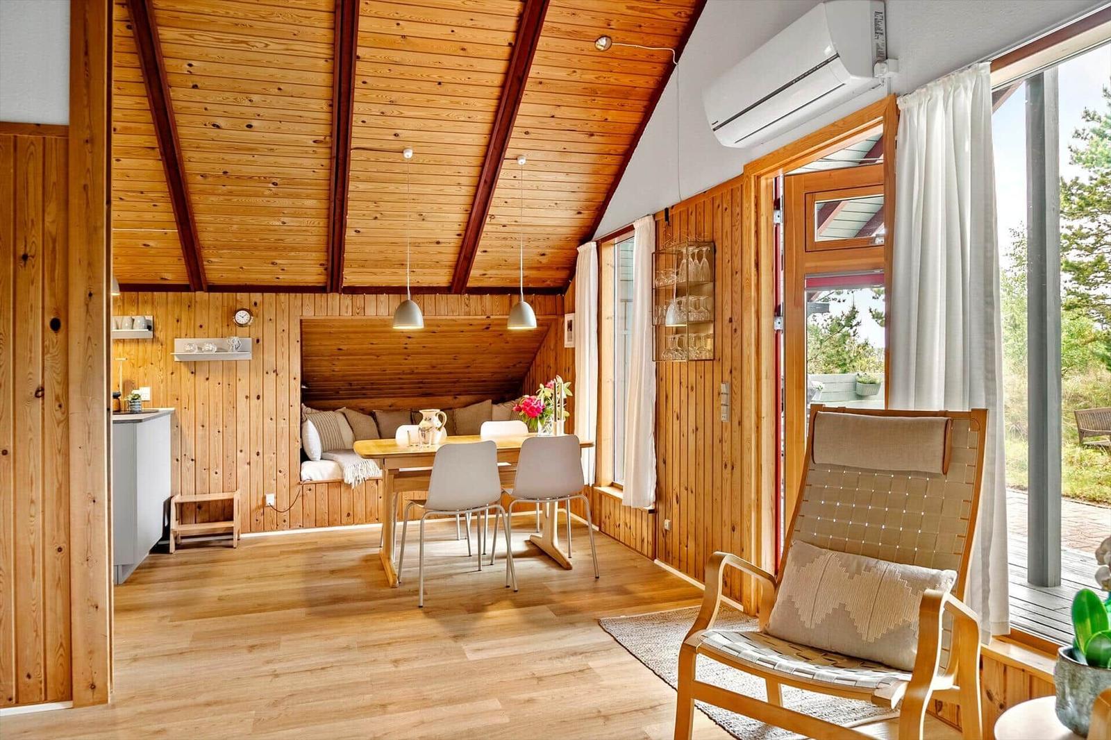 Wohnraum mit Holzwand, Esstisch, Sitzbank und Sessel. Durchgang zur Küche. Große Fenster mit Blick.