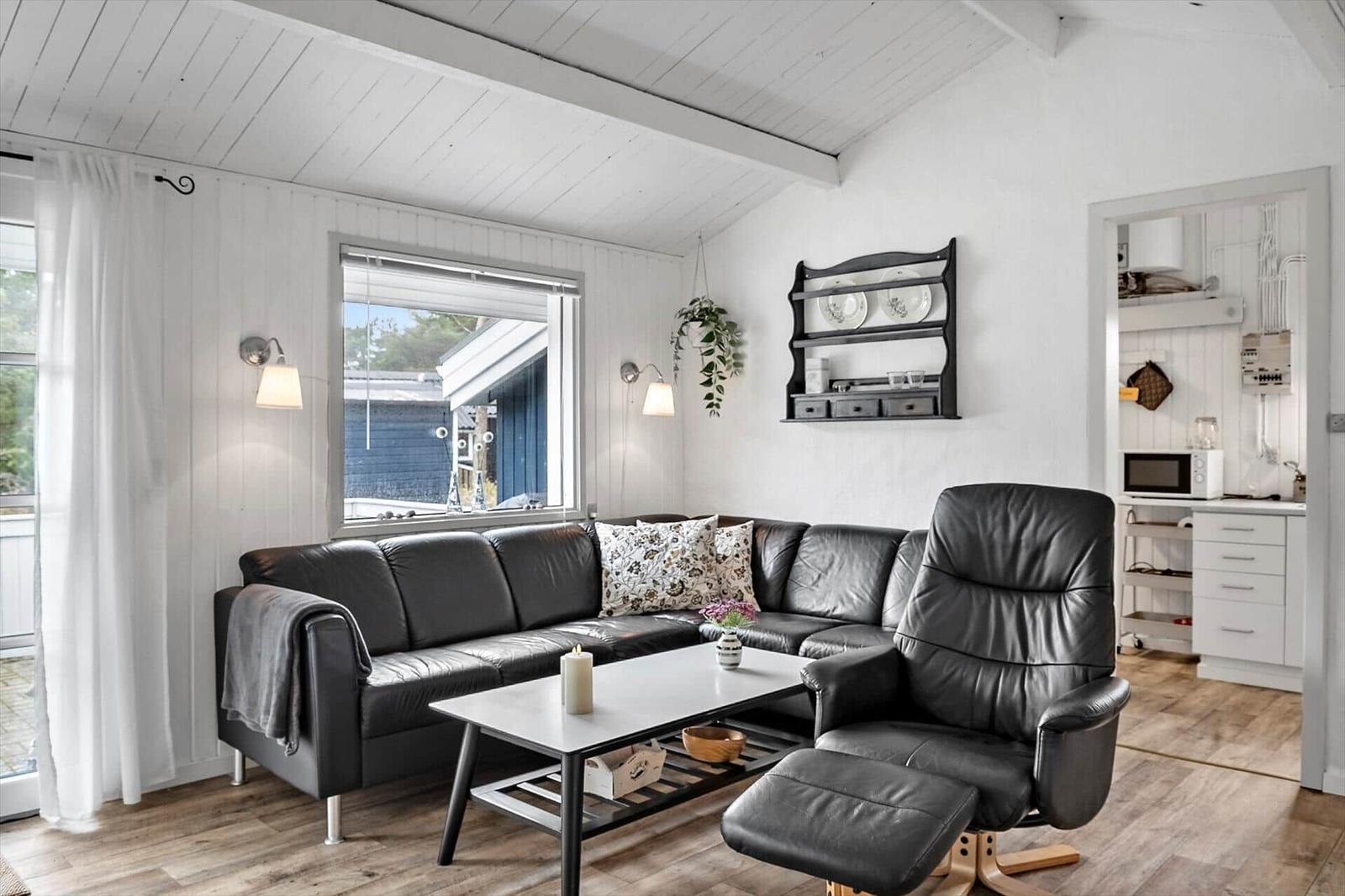 Wohnzimmer mit Sofa, Sessel und Tisch. Küche im Hintergrund. Weiße Wände und Holzboden.
