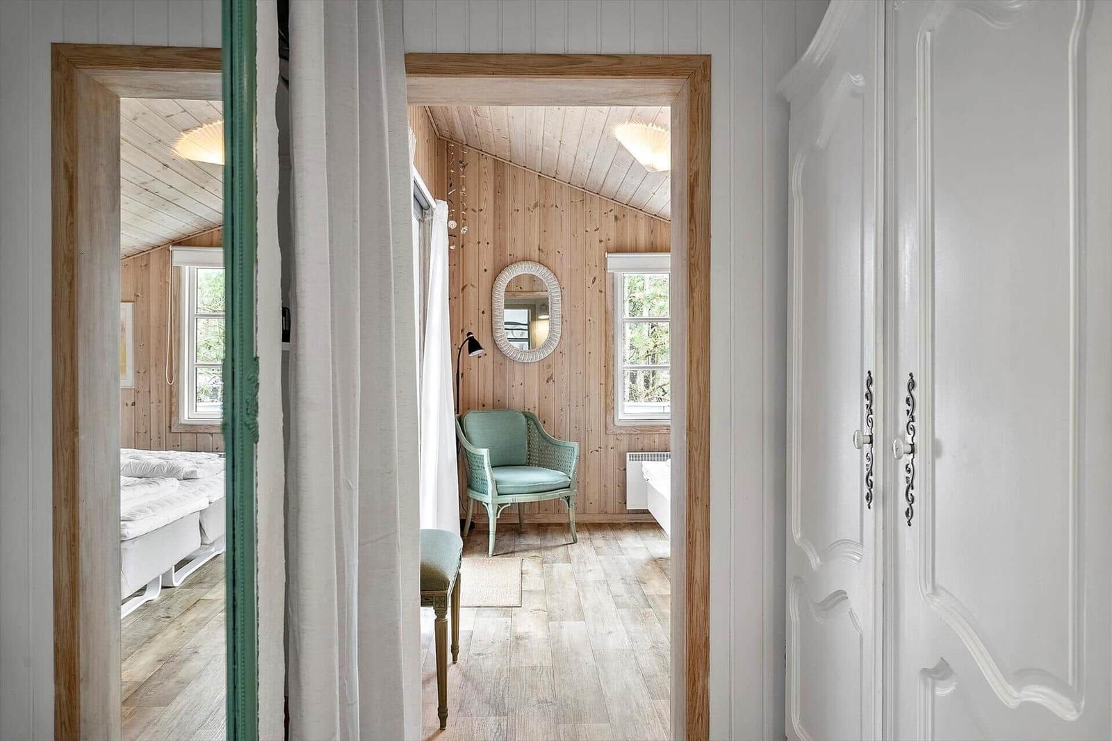 Schlafzimmer mit Holzwand, Bett und Stuhl. Blick in einen Flur mit Spiegel und Fenster.