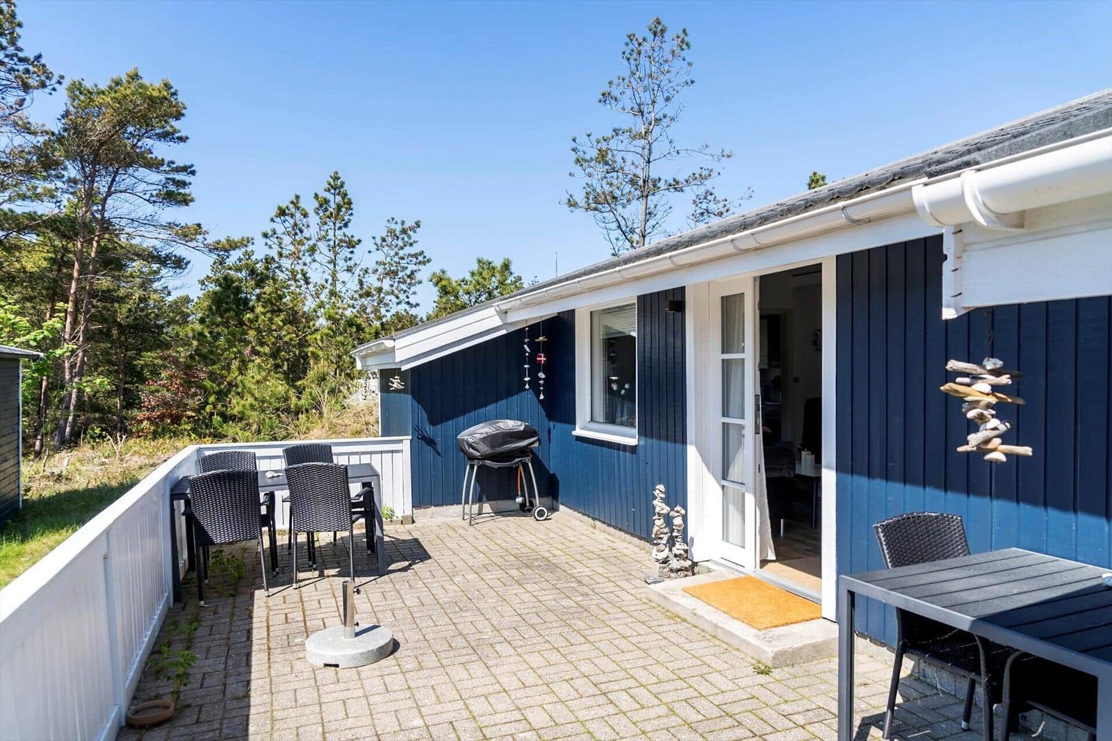 Blauer Ferienhaus mit Terrasse, Grill und Sitzgelegenheiten. Umgeben von Bäumen und klarem Himmel.
