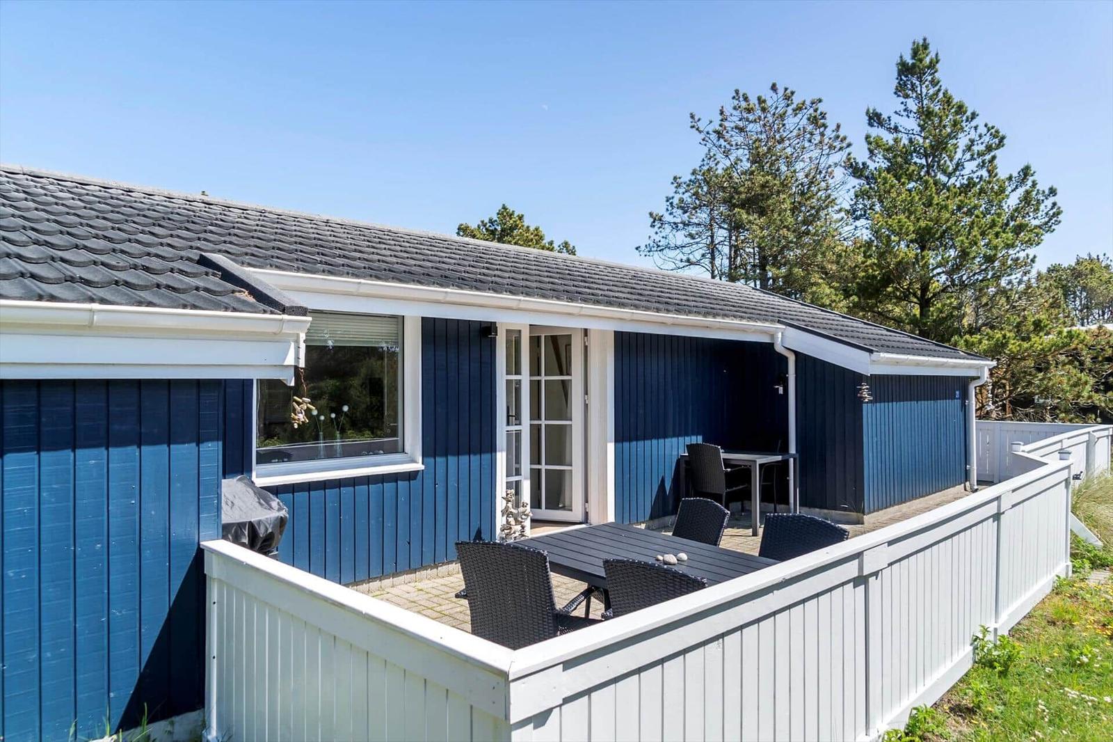 Ein blaues Ferienhaus mit Terrasse, Tisch und Stühlen. Hinterhof mit weißem Zaun und Bäumen.