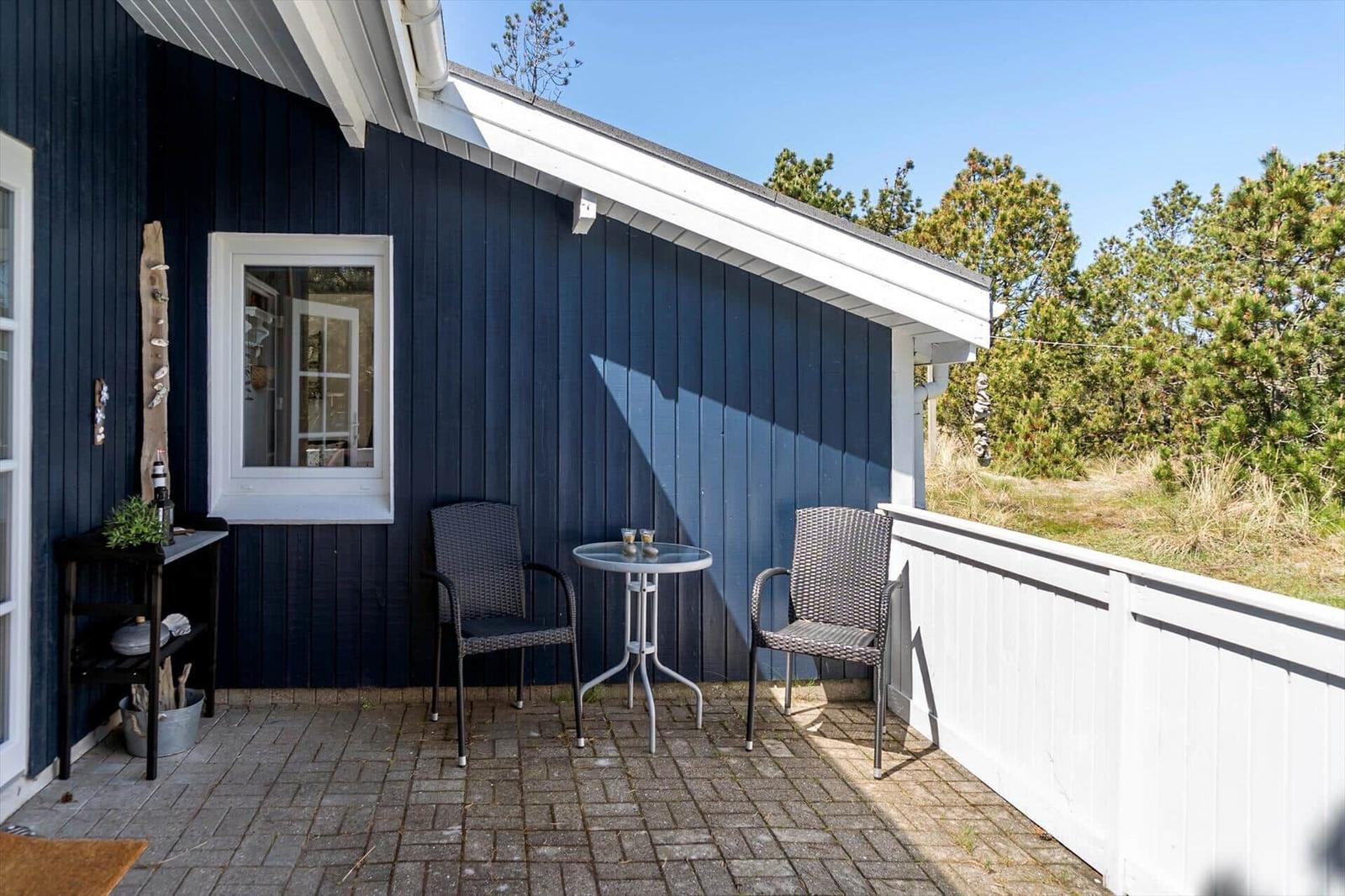 Terrasse mit Tisch und zwei Stühlen vor blauem Holzhaus mit weißem Fenster und Zaun.
