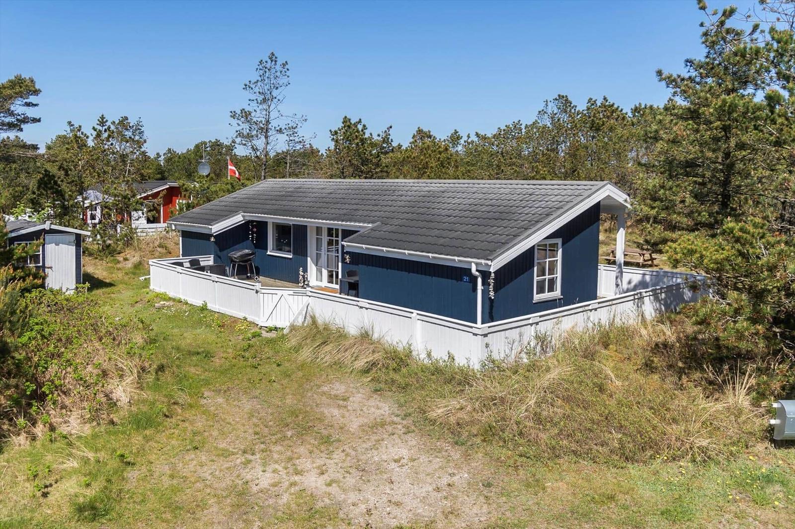 Blau gestrichenes Haus mit weißem Zaun und Terrasse in einer grünen Umgebung.