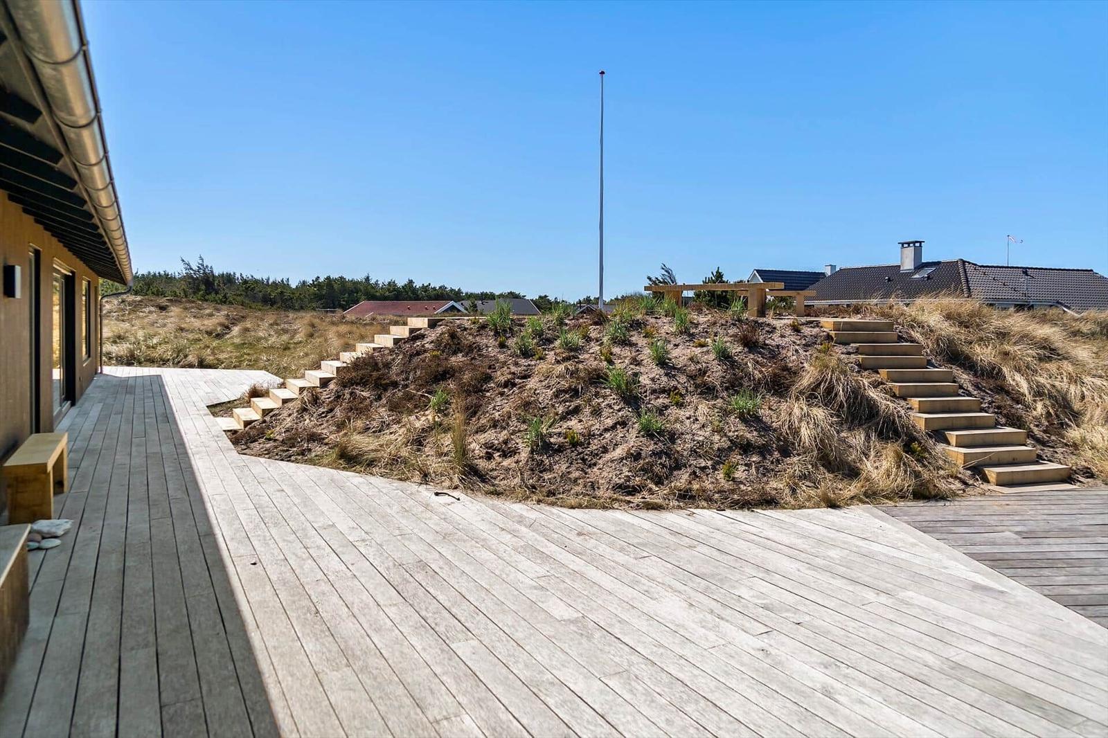Holzterrasse mit Steigstufen zu einer Dünenlandschaft mit Blick auf Häuser und einen Mast.