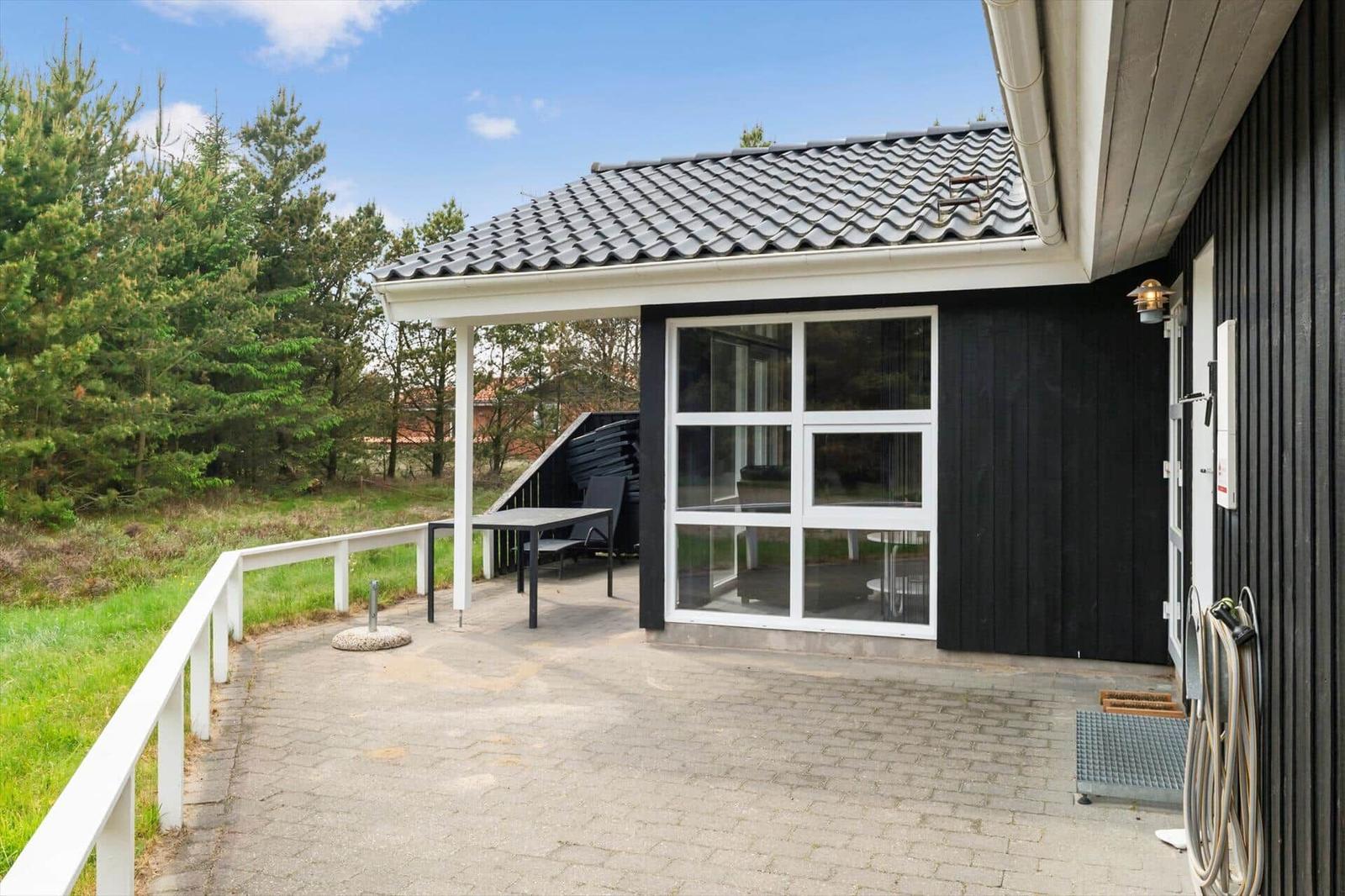 Schwarzes Haus mit Terrasse, Tisch und Stühlen. Glasfront und Holzverkleidung. Hinterhof mit Bäumen.