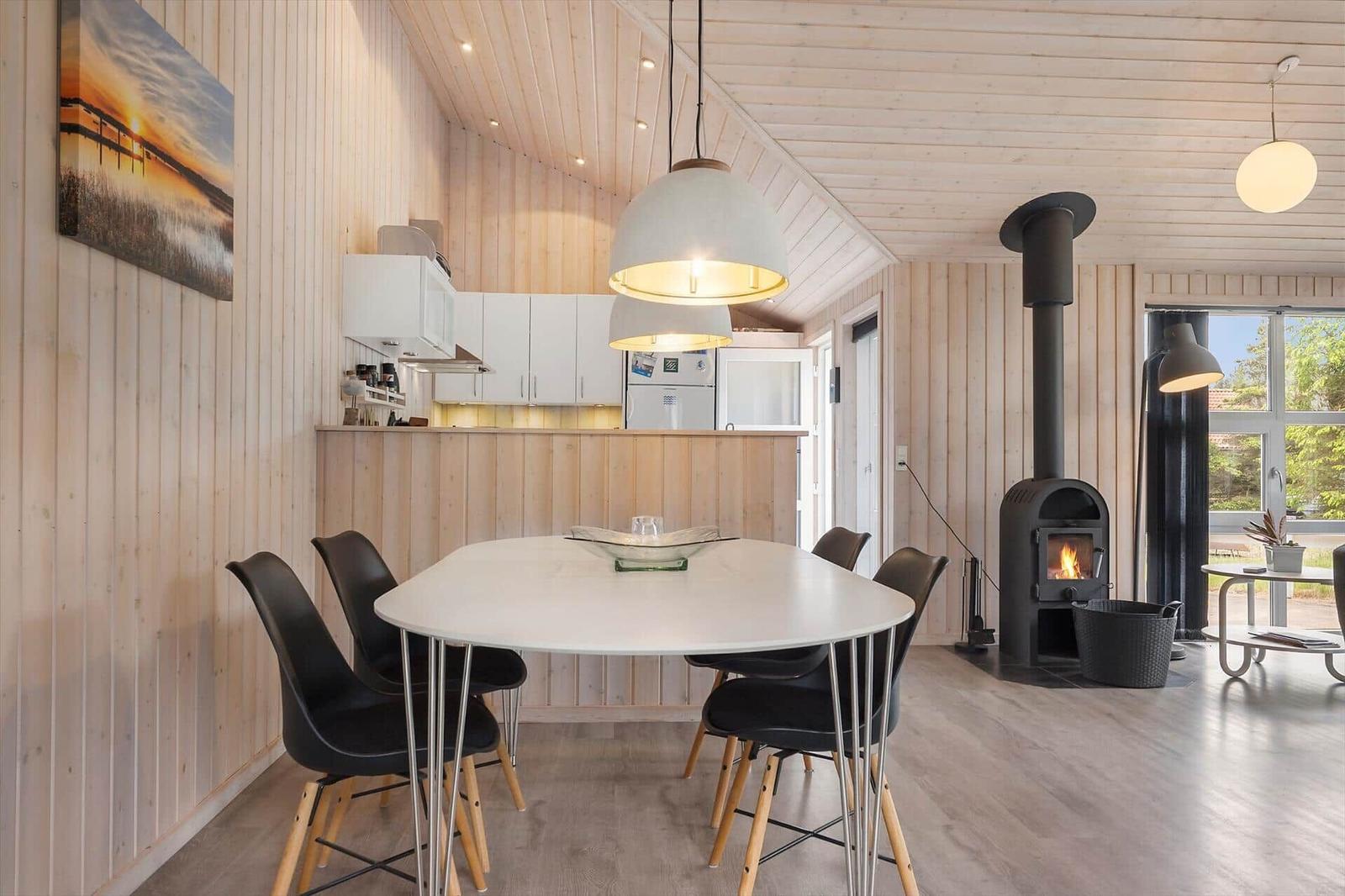 Essbereich mit Tisch, Stühlen und Kaminofen. Küche mit weißen Schränken. Holzwände und Decke.
