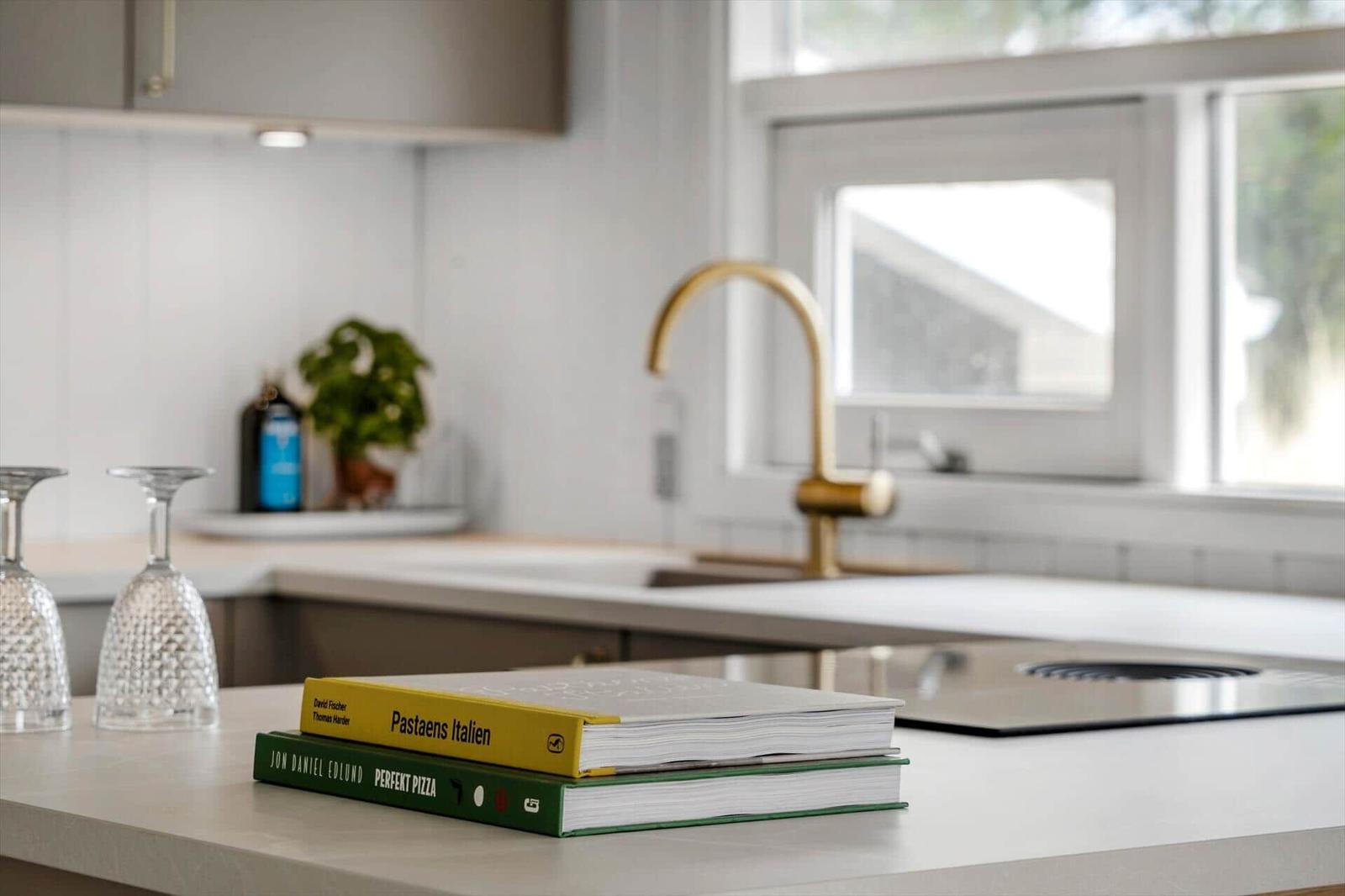 Kücheninsel mit Kochbuch und Gläsern. Hintergrund: goldener Wasserhahn und Fenster.