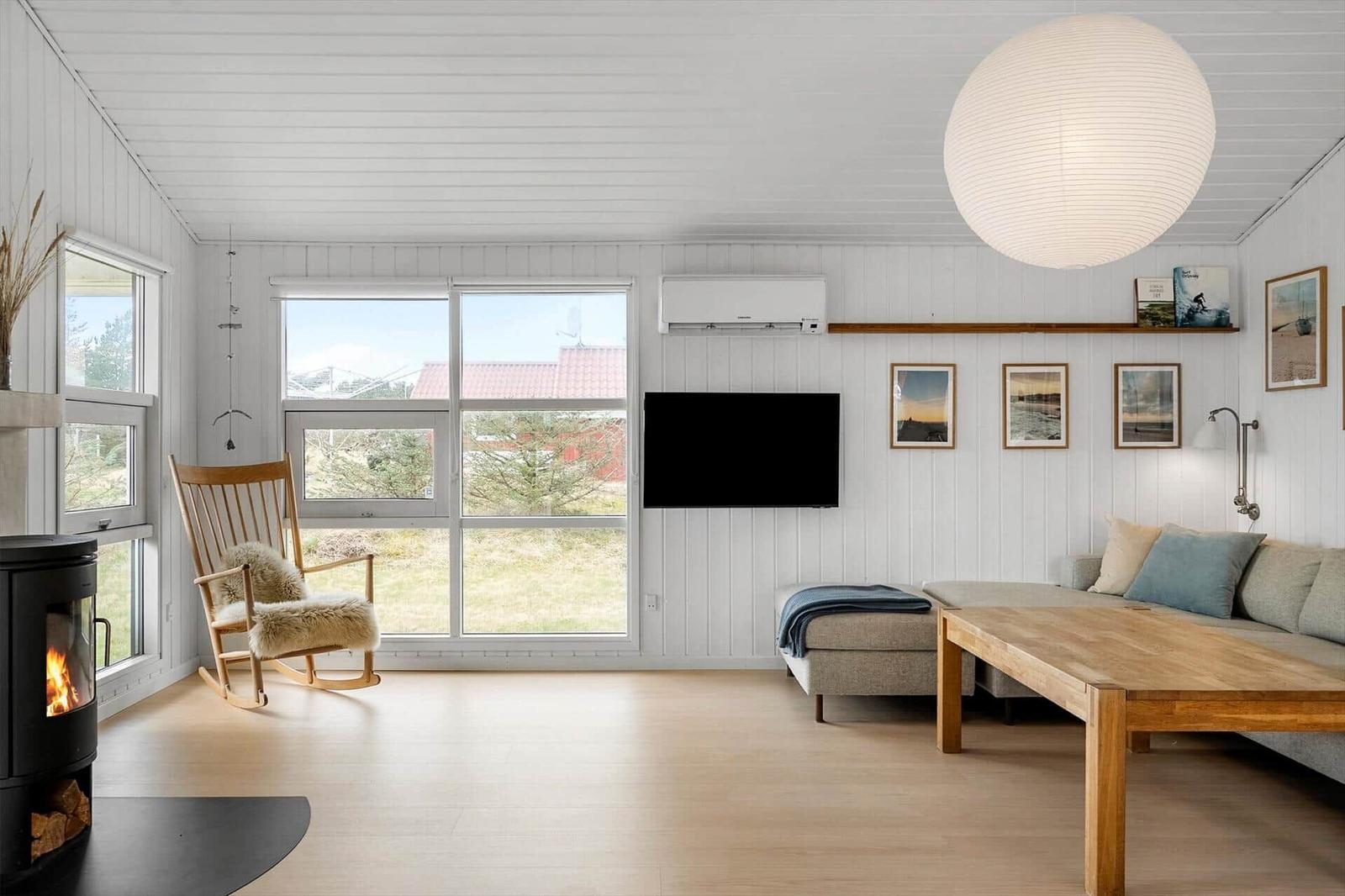 Wohnzimmer mit Holzfußboden, Fernseher, Sitzbank und Schaukelstuhl. Große Fenster und Deckenlampe.
