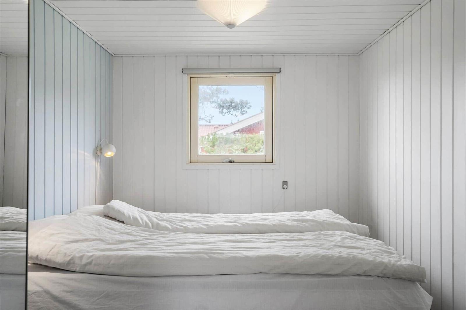 Schlafzimmer mit weißem Bett und Fenster. Wände sind weiß verkleidet.