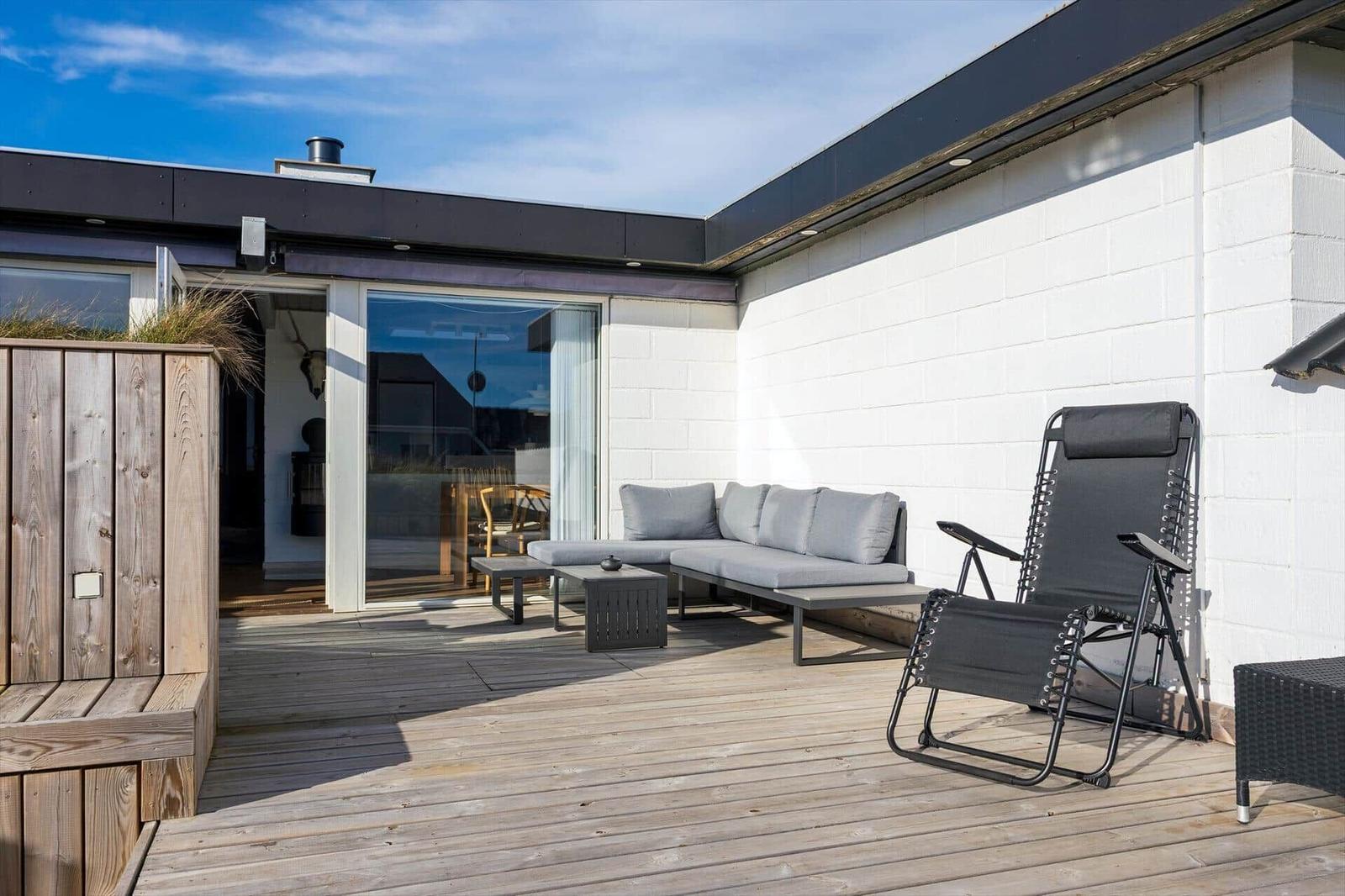 Terrasse mit Sofa, Liegestuhl und Holzboden. Glasfront führt ins Haus.