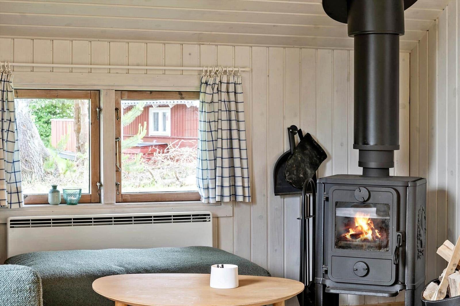 Room with wood stove, window with curtains and table. Outside, a red building is visible.