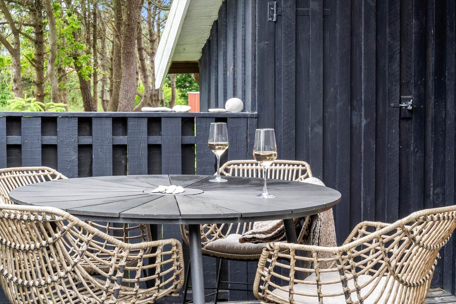 Round table with chairs and wine glasses on a terrace in front of a dark wooden house.