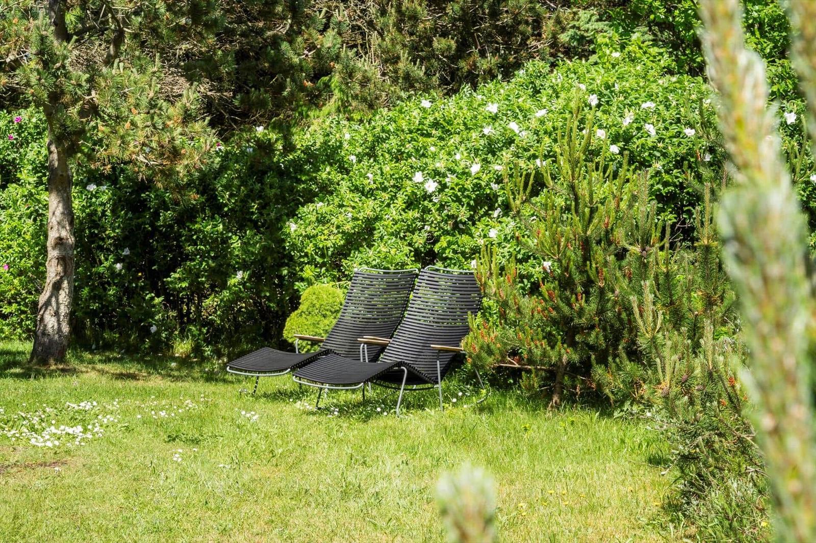 Two black lounge chairs are on a green lawn between shrubs and trees.