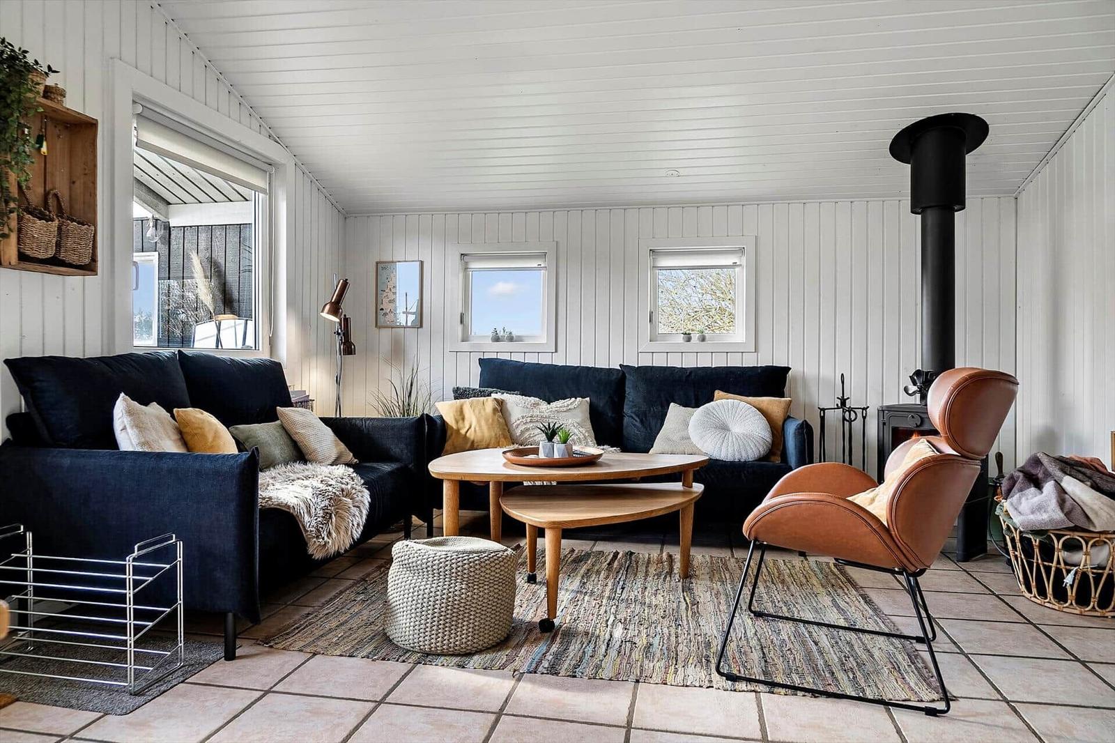 Wohnzimmer mit zwei dunklen Sofas, Holztisch und Ledersessel. Weiße Holzwände und Kachelboden.
