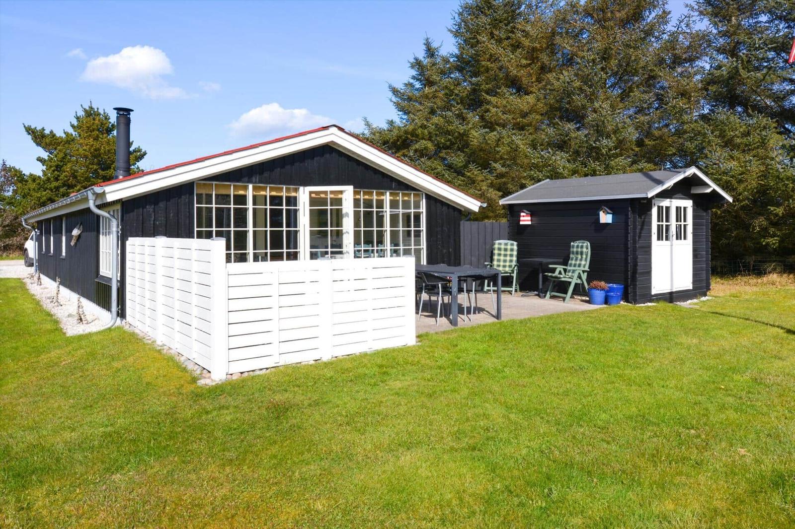 Schwarze Holzhütte mit weißen Fenstern und Terrasse. Grünfläche und Bäume umgeben das Grundstück.