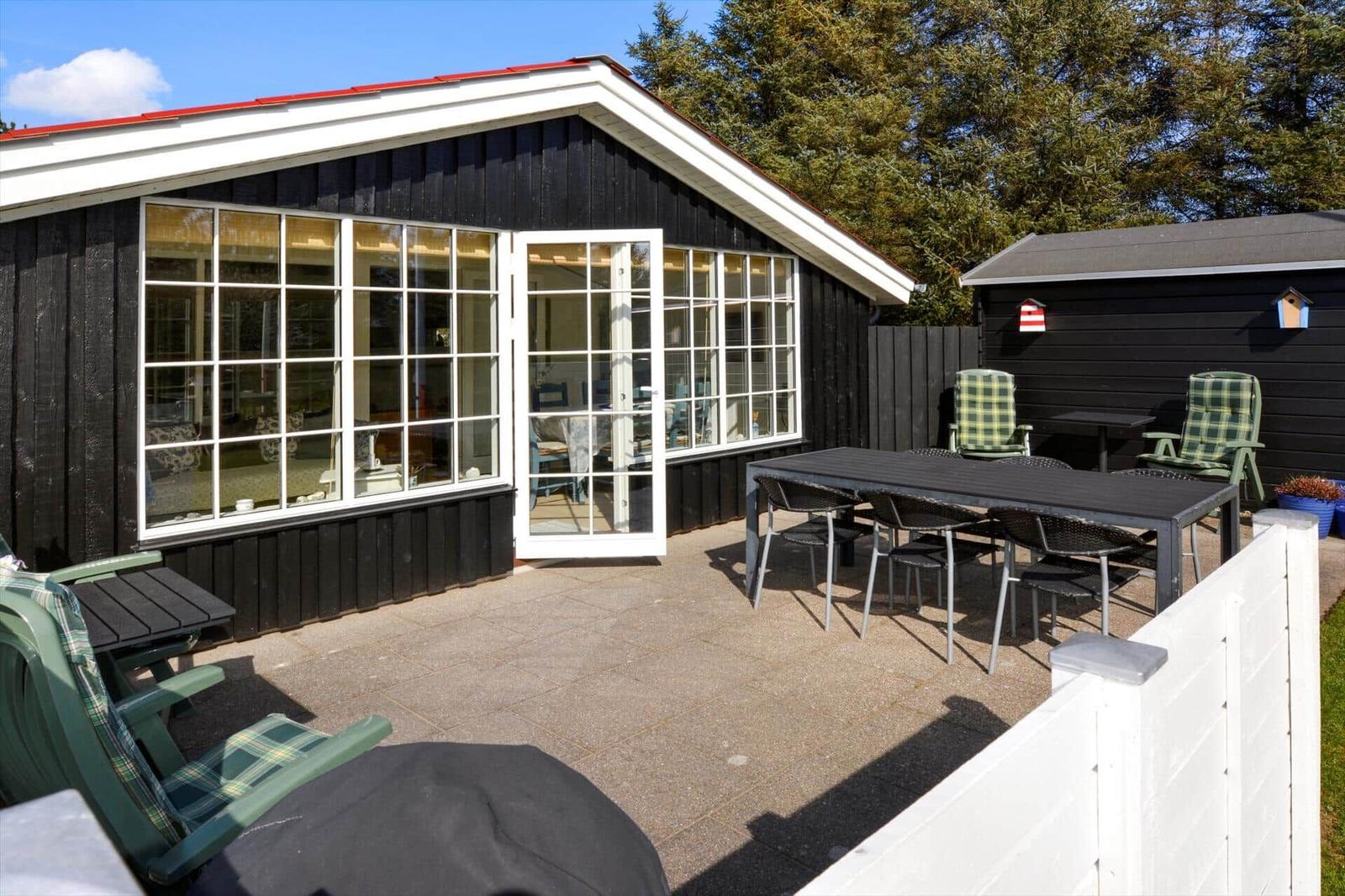 Haus mit Terrasse, Tisch, Stühlen und Gartenmöbeln. Schwarze Fassade, weiße Fenster und roter Dachrand.