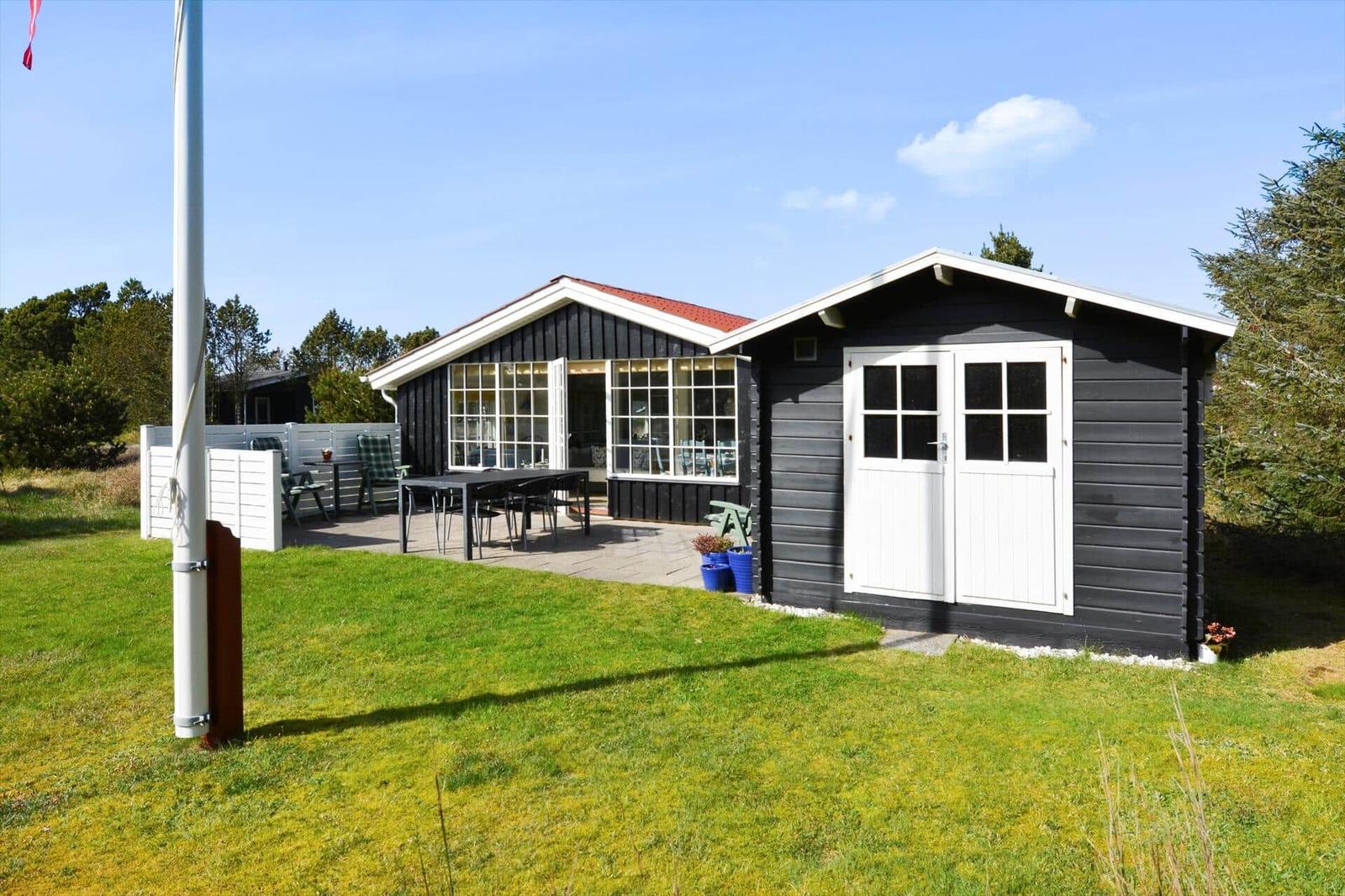 Schwarze Holzhütte mit weißer Tür und Terrasse. Grünflächen und Bäume umgeben das Haus.