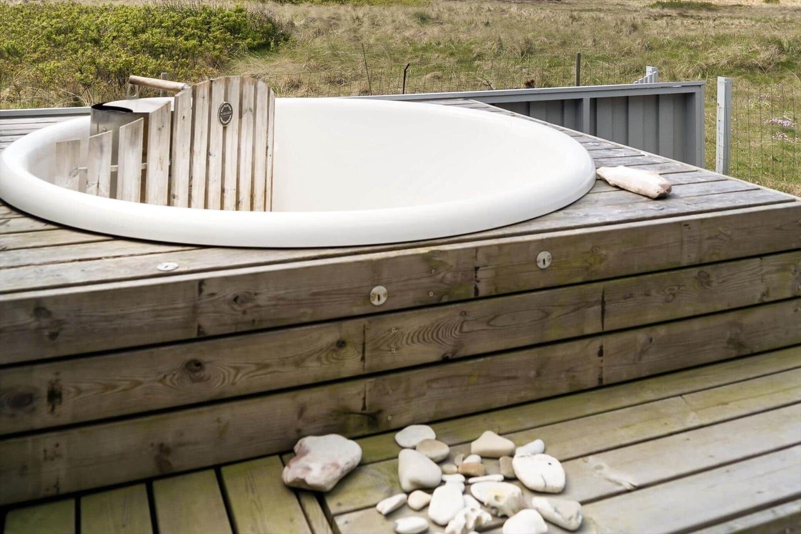 Runde Whirlpool-Badewanne auf Holzterrasse mit Steinen und Blick auf Grasland.