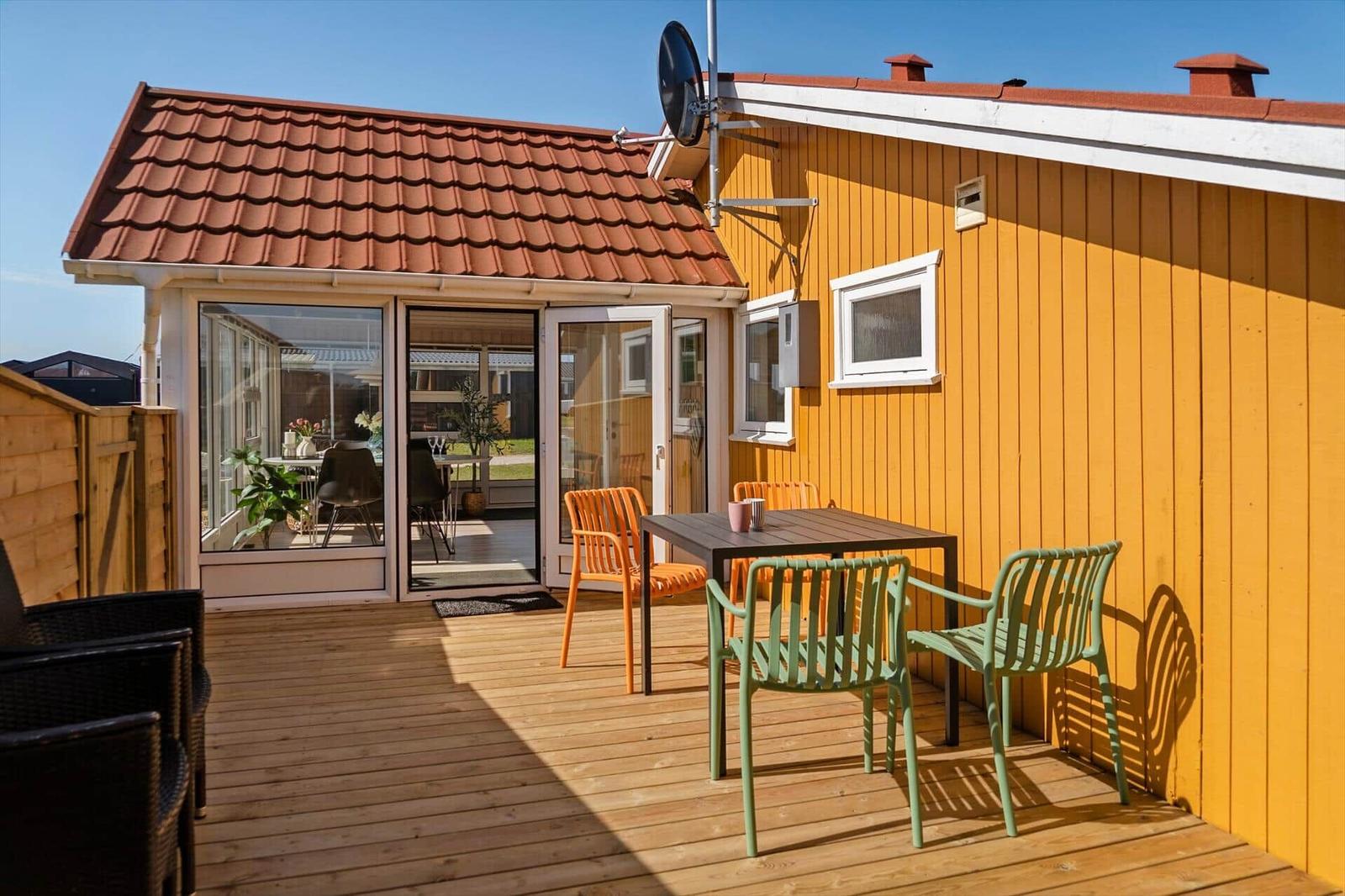 Terrasse mit Tisch und Stühlen vor gelbem Haus mit Glasfront und Dach.