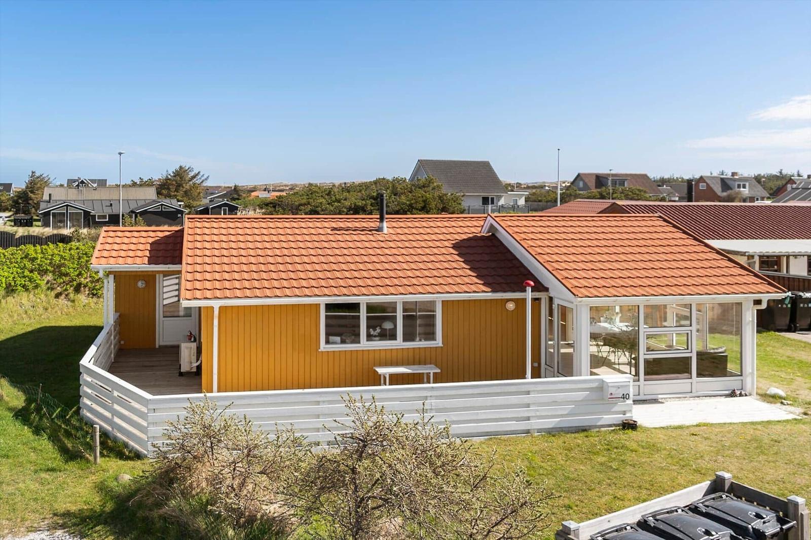 Ein gelbes Haus mit rotem Dach und weißer Veranda. Ein Garten und Nachbarhäuser sind sichtbar.