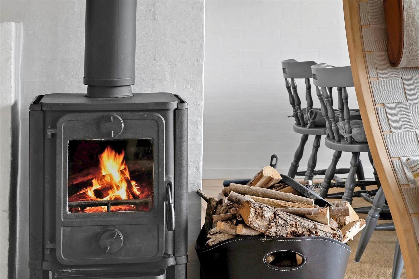 Holzofen mit brennendem Feuer, daneben Stapel Brennholz und Stühle.