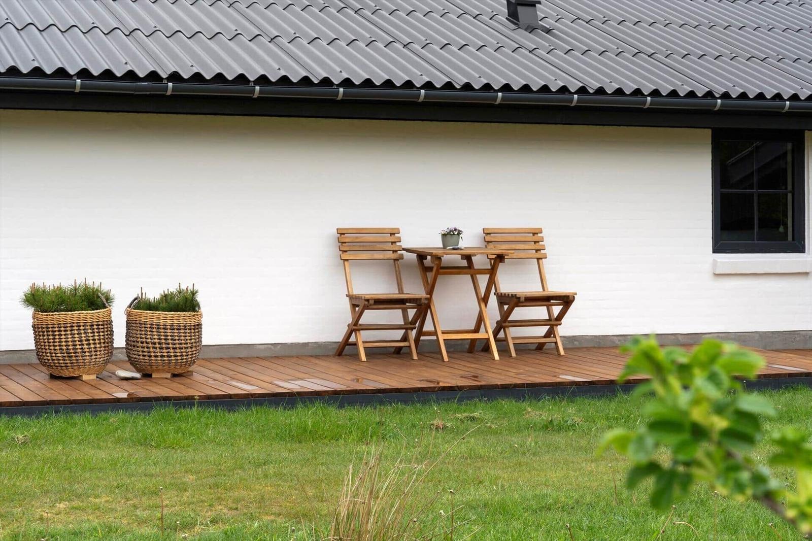 Terrasse mit Holztisch, zwei Stühlen und zwei Körben mit Pflanzen an weißer Wand.