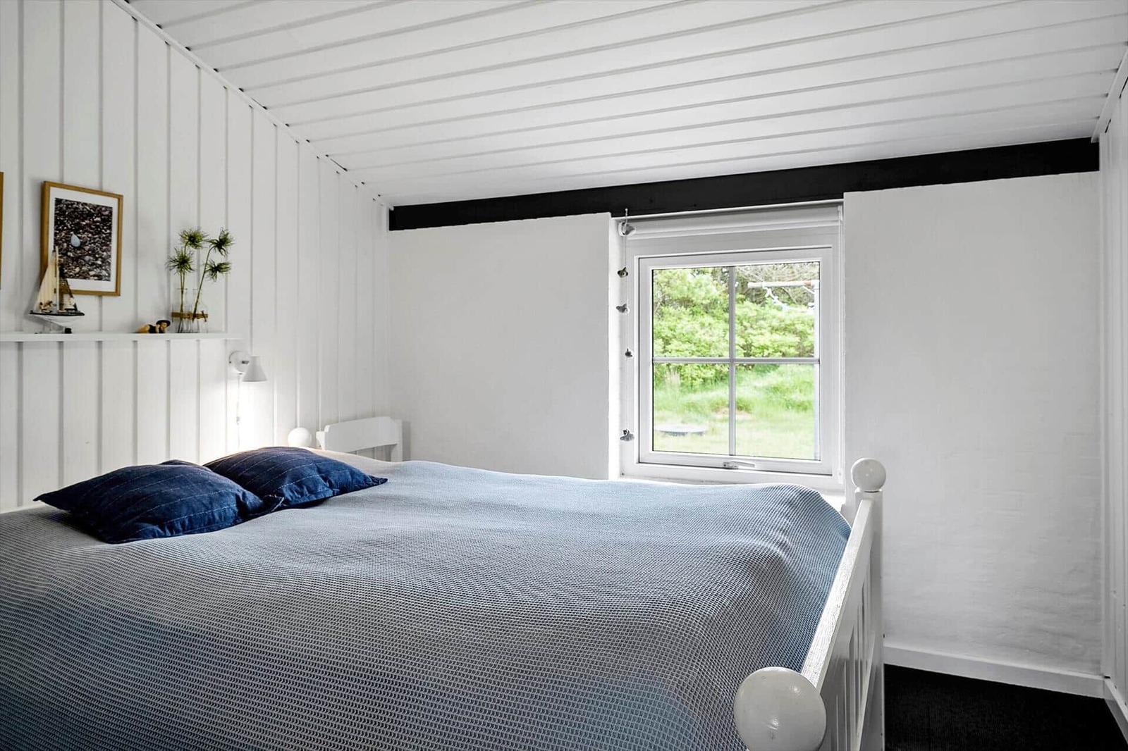 Schlafzimmer mit weißer Holzwand, Bett mit blauer Bettwäsche und Fenster zum Garten.