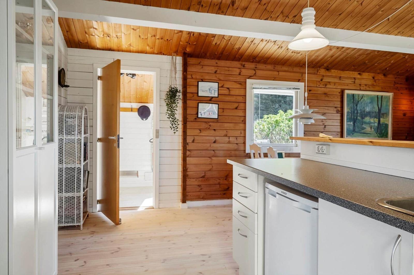 Küche mit Arbeitsplatte, Spüle und Waschmaschine. Holzwände und -decke. Tür führt in einen Raum.