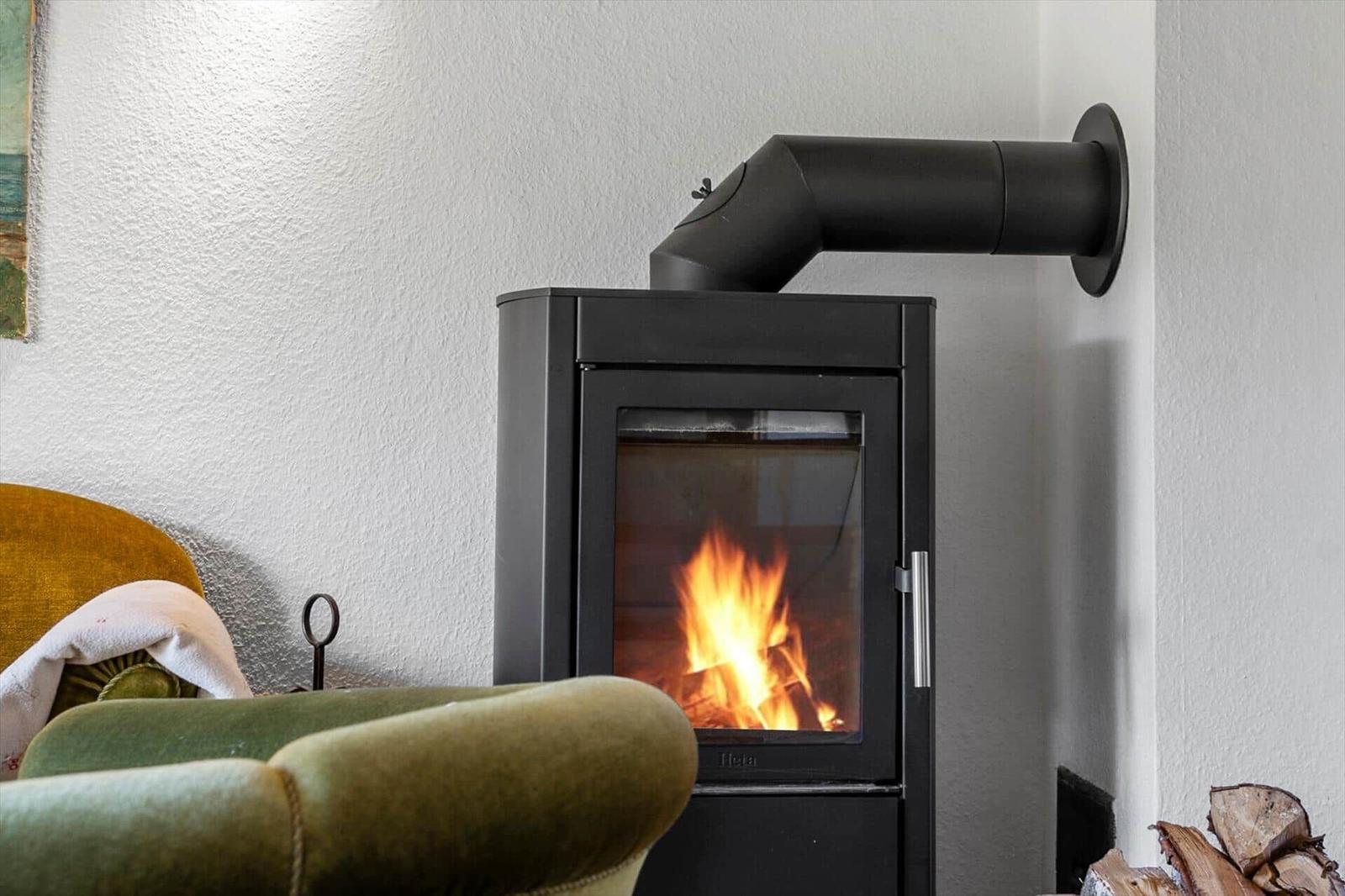 A wood-burning stove with burning logs, next to a green armchair and a stack of firewood.